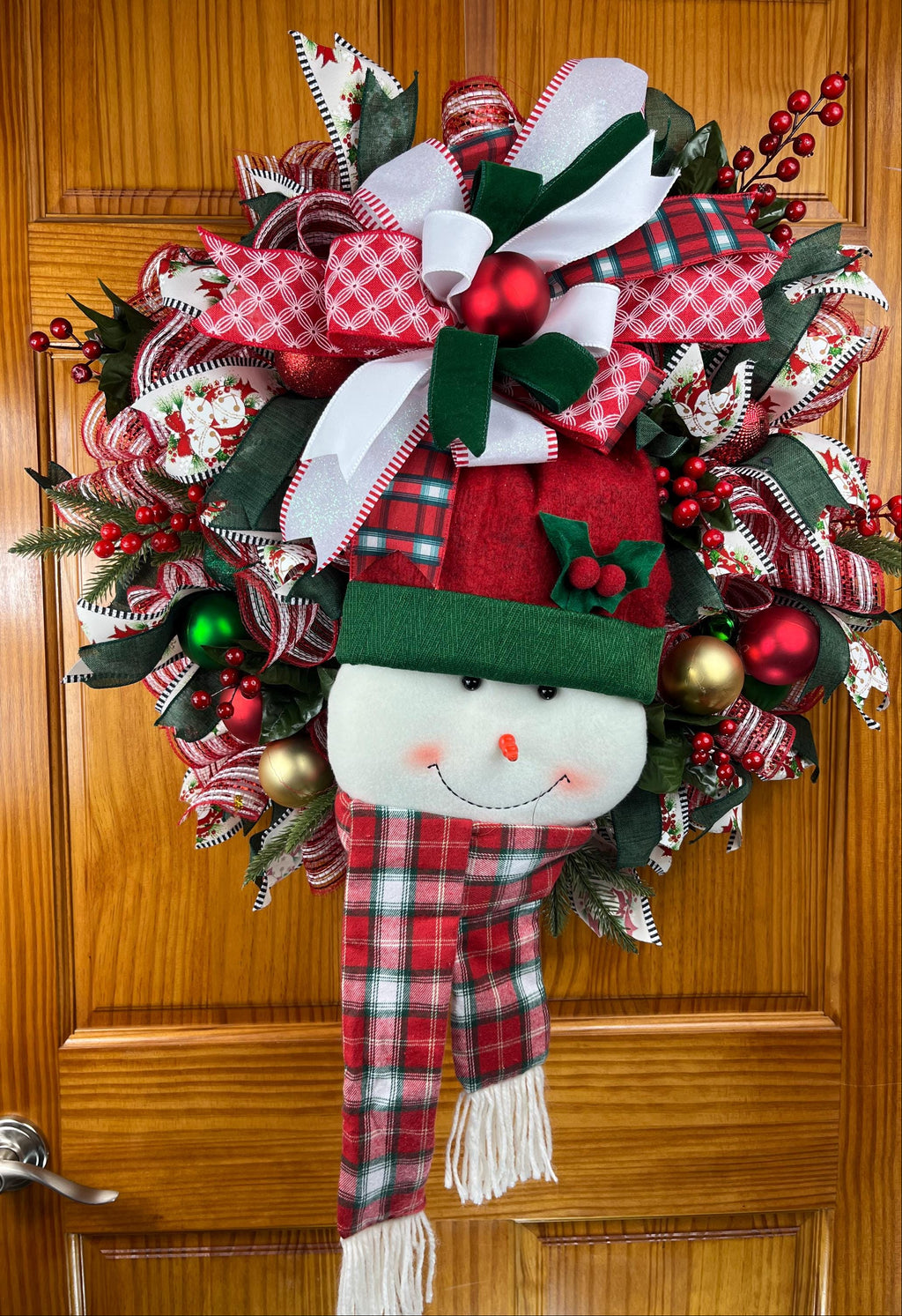 A festive Christmas wreath hanging on a wooden door, featuring a plush snowman face with a green hat, red plaid scarf, red berries, green and white ribbons, and holiday ornaments.