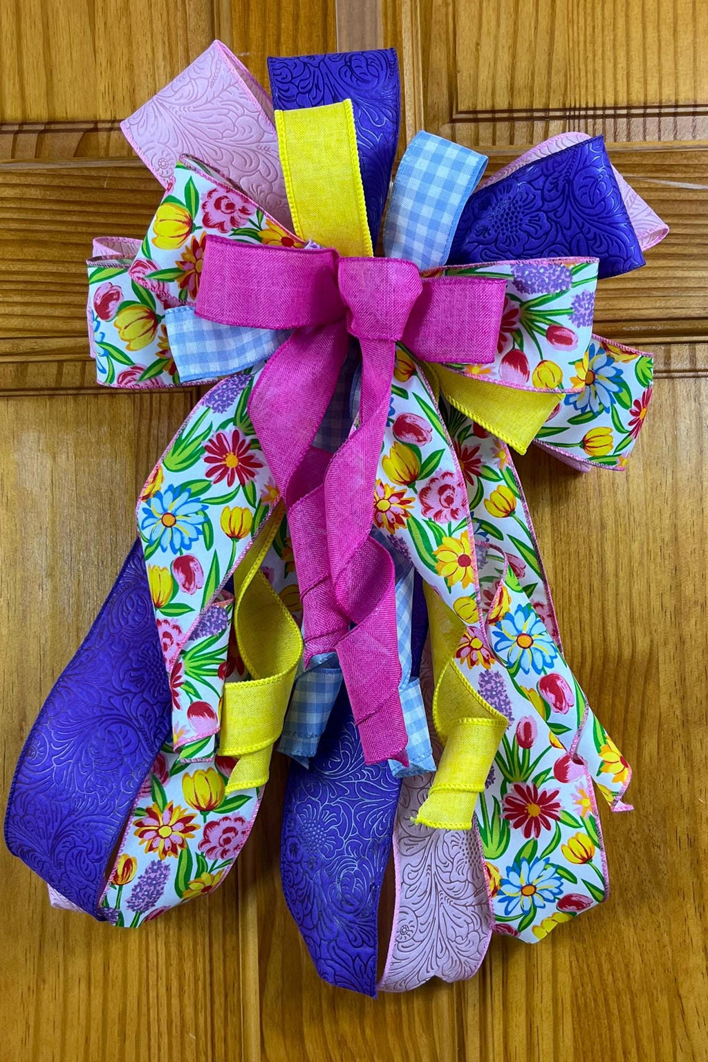 A colorful handmade fabric ribbon bow with a magenta center and loops of purple, yellow, floral, and gingham ribbon, hung on a brown wooden door.