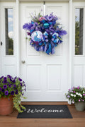 A decorative wreath hanging on a wooden door, featuring blue and purple mesh ribbons, faux flowers, feathers, and a peacock-themed ornament.