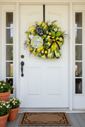 A decorative spring wreath hanging on a brown wooden door. The wreath features yellow and green mesh ribbon, white daisies, yellow pom-poms, a black and white plaid bow, and a round sign that reads 'Home with Love' in script over a lemon pattern.