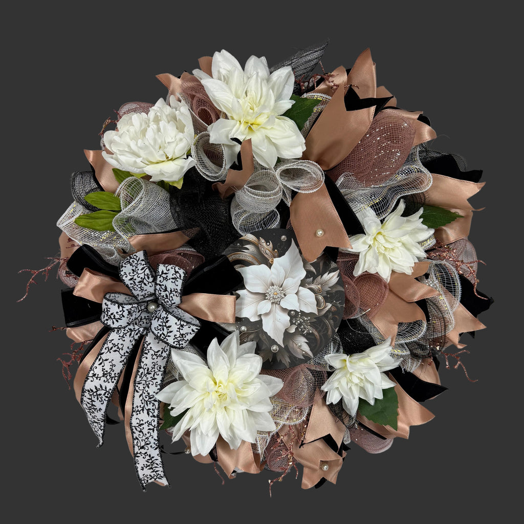 A decorative mesh wreath featuring white faux dahlia flowers, layered with black, taupe, and silver mesh ribbons, and a black-and-white patterned bow, displayed against a white fabric backdrop.