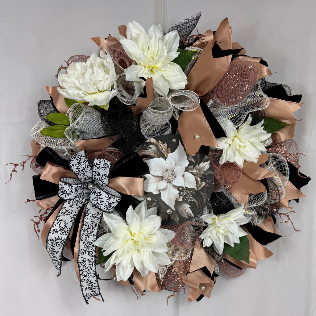 A decorative autumn wreath featuring white faux dahlia flowers, layered with metallic bronze mesh, black mesh, sheer gray mesh, and a black-and-white patterned ribbon bow, displayed against a white fabric backdrop.