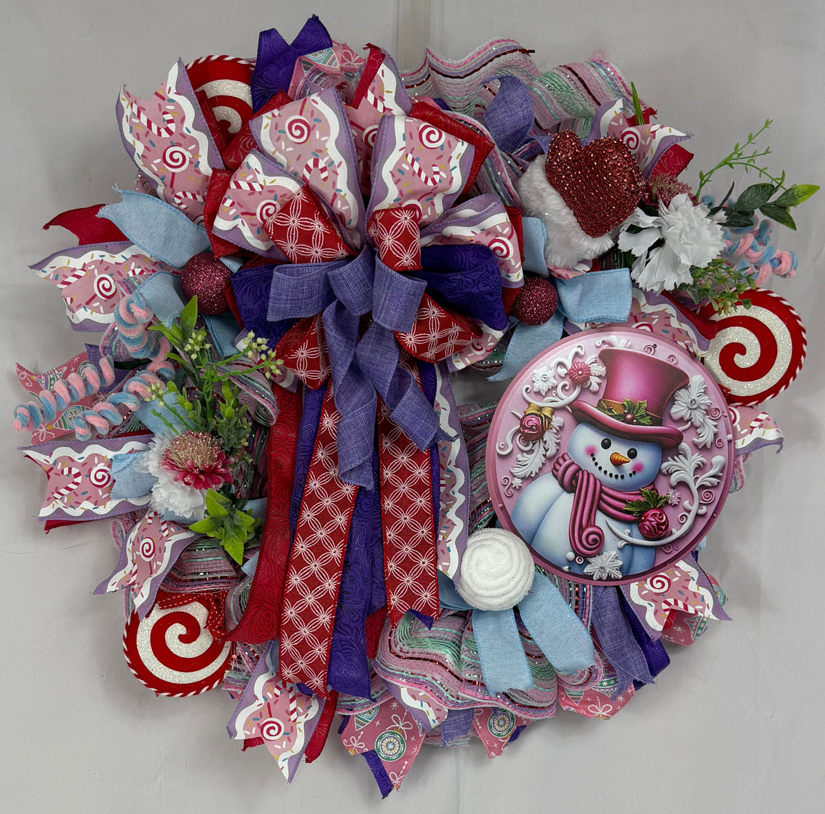 A festive Christmas wreath made of layered red, white, and purple ribbons, candy cane accents, faux greenery, and a round snowman ornament, hanging against a white fabric backdrop.