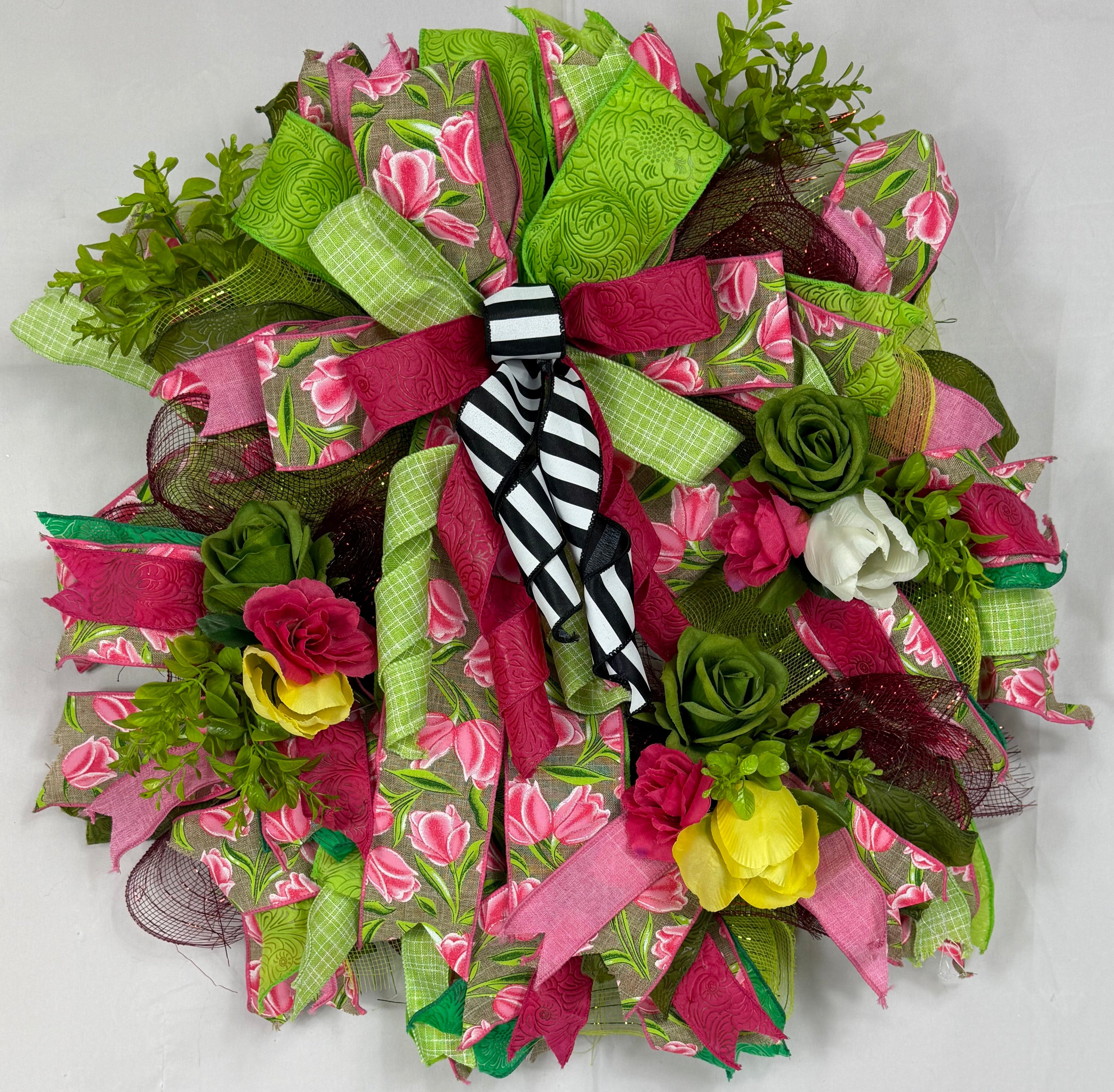 A decorative mesh wreath featuring pink and green floral patterns, green mesh ribbon, faux roses, and a black-and-white striped bow, displayed against a white fabric backdrop.