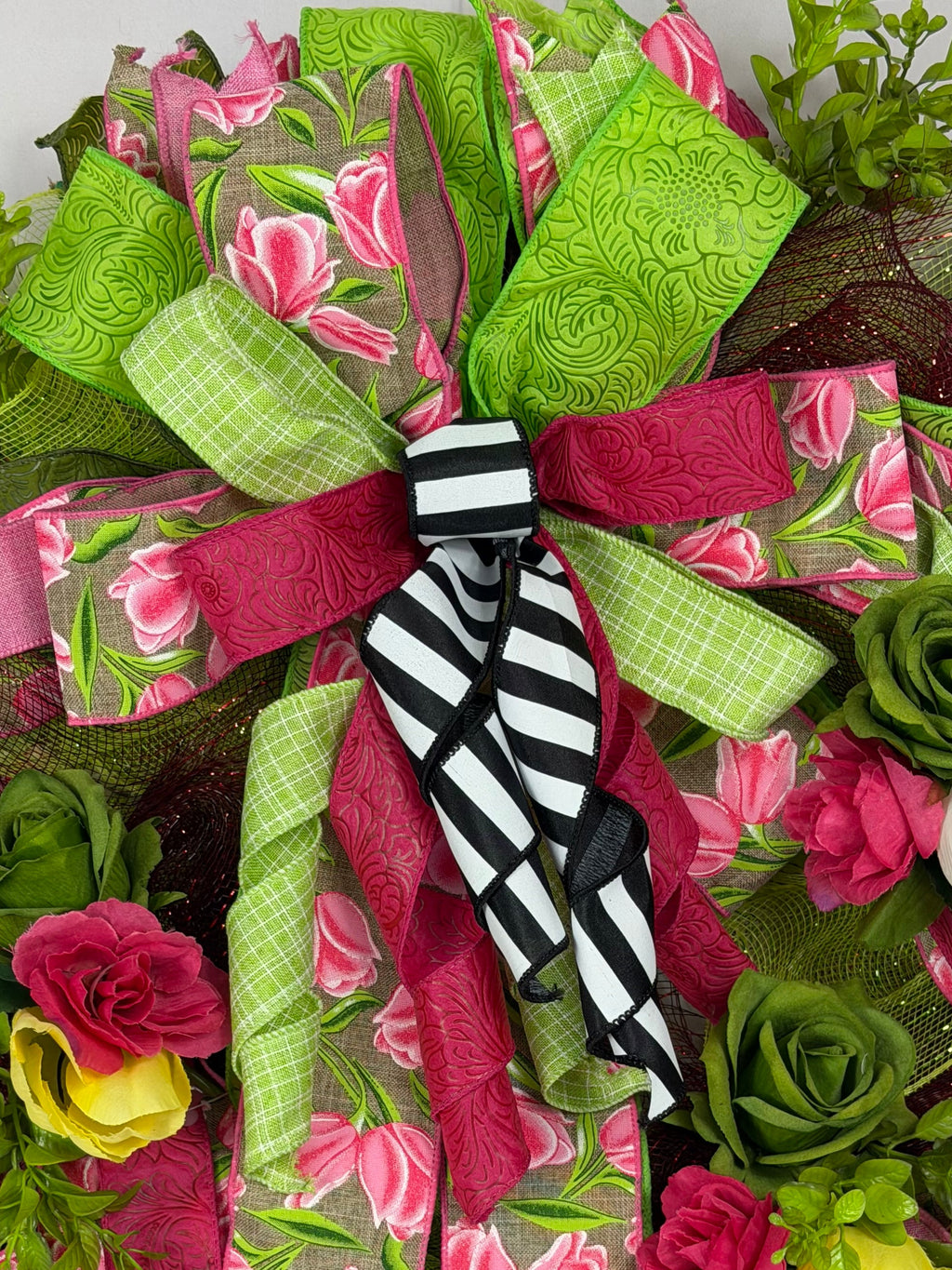 A decorative mesh wreath with layered green and pink floral ribbon, faux roses, greenery, and a black-and-white striped bow at the center, photographed against a plain light background.