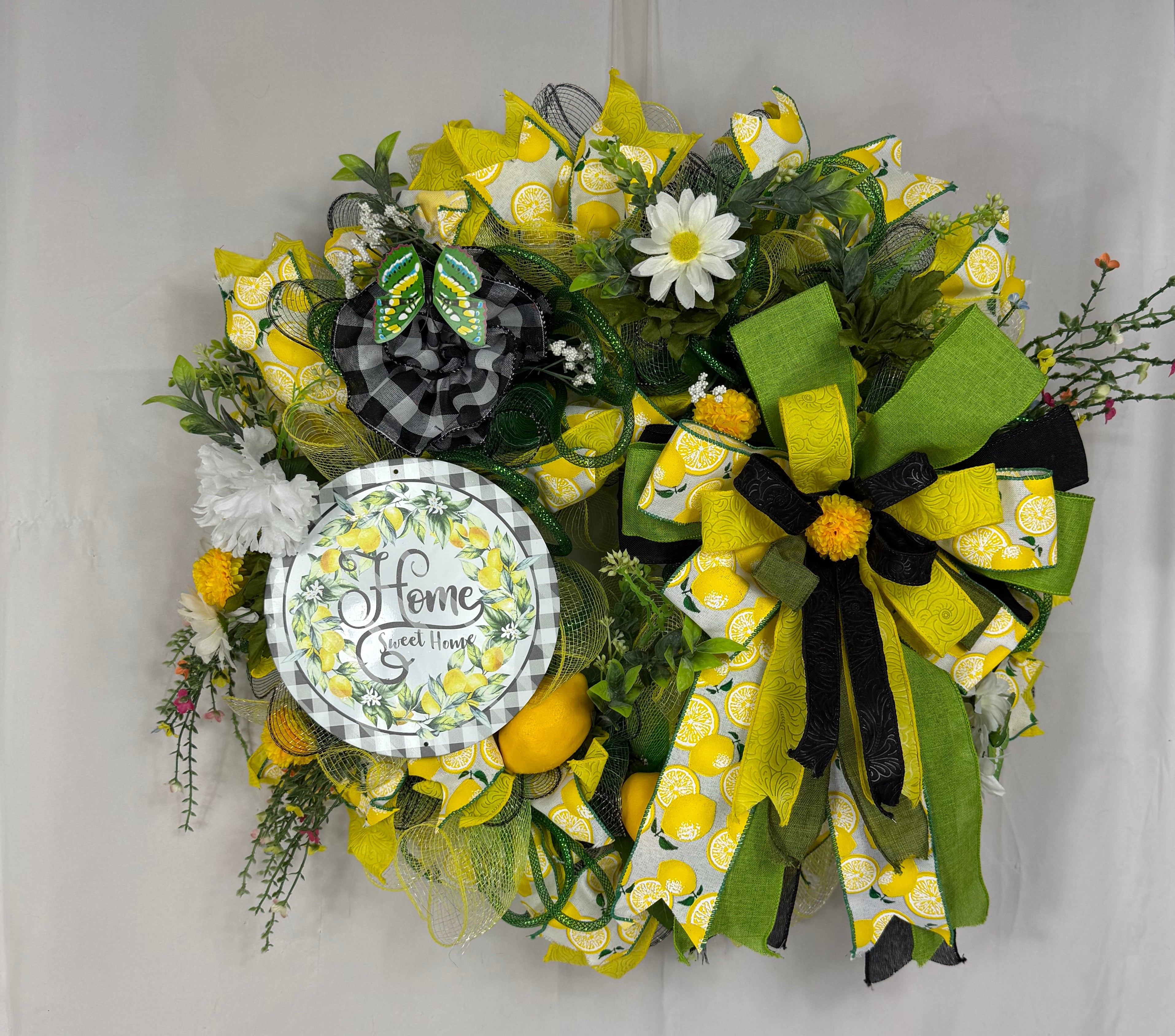 A decorative floral wreath hanging on a white door. The wreath features yellow and green patterned ribbons, black and yellow bows, green mesh, faux daisies, small yellow pom-poms, and a round white sign that reads 'Home Sweet Home'.