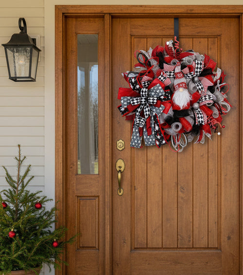 A festive Christmas mesh wreath in red, black, and white, featuring large patterned bows, silver glitter accents, a small white beard gnome figure, and red and white striped ribbons, displayed on a white paneled door.